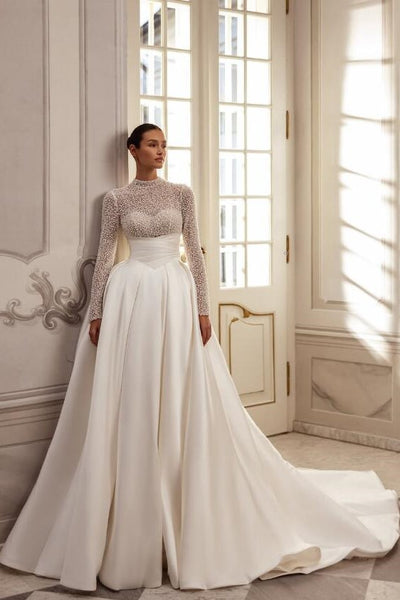 Woman in a white wedding dress with long sleeves standing in a room with large windows.